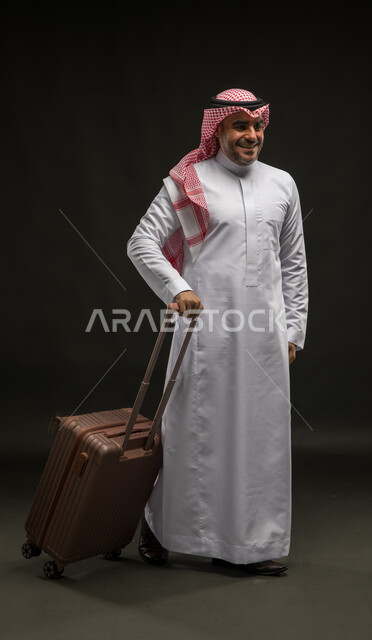 ايماءات النشاط والحيوية و الاستعداد لقضاء رحلة صيفية ممتعة, بورتريه لرجل عربي سعودي خليجي مبتسم يرتدي الشماغ والثوب التقليدي و يمسك بيده حقيبة السفر, الشعور بالحماس والفرح، صورة الجسم بالطول الكامل, خلفية سوداء.