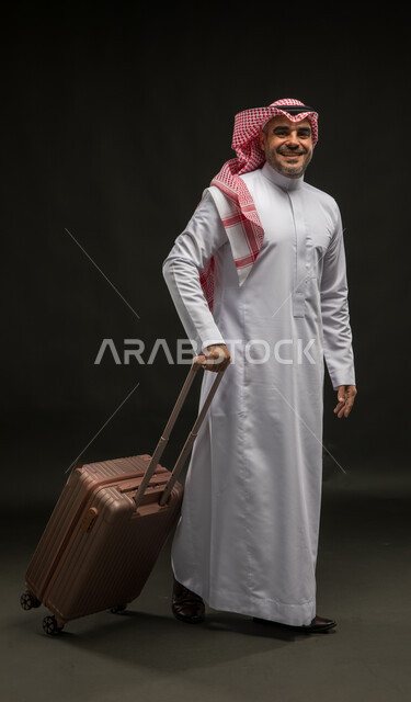 النظر إلى الكاميرا بإيماءات النشاط والحيوية و الاستعداد لقضاء رحلة صيفية ممتعة, بورتريه لرجل عربي سعودي خليجي مبتسم يرتدي الشماغ والثوب التقليدي و يمسك بيده حقيبة السفر, الشعور بالحماس والفرح، صورة الجسم بالطول الكامل, خلفية سوداء.