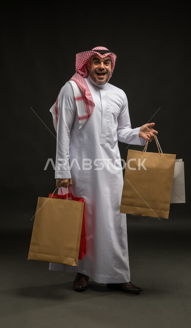 الاستفادة من العروضات والتخفيضات، المتعة في التسوق واقتناء المستلزمات والهدايا، بورتريه لرجل عربي خليجي سعودي مبتسم يرتدي الشماغ والثوب التقليدي يحمل بيده أكياس ورقية ملونة بإيماءات الدهشة والذهول، صورة الجسم بالطول الكامل، خلفية سوداء