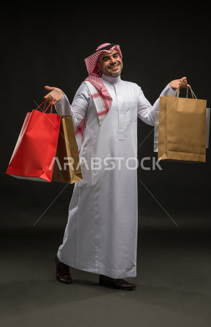 المتعة في التسوق واقتناء المستلزمات والهدايا، الاستفادة من العروضات والتخفيضات، بورتريه لرجل عربي خليجي سعودي مبتسم يرتدي الشماغ والثوب التقليدي يحمل بيده أكياس ورقية ملونة بإيماءات السرور والسعادة، صورة الجسم بالطول الكامل، خلفية سوداء