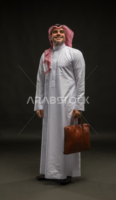 الذهاب إلى دوام العمل بنشاط, الشعور بالسرور لتحقيق أهداف الشركة, بورتريه لرجل عربي سعودي خليجي مبتسم يرتدي الشماغ والثوب التقليدي يحمل حقيبة يد, الاهتمام بالأناقة والمظهر الخارجي، صورة الجسم بالكامل, خلفية سوداء
