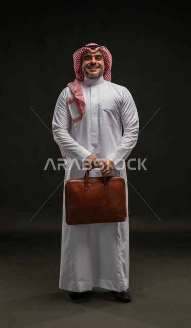 Standing upright with gestures of self-confidence, going to work energetically, portrait of a smiling Saudi Arabian Gulf man wearing a shemagh and traditional dress looking at the camera holding a handbag, attention to elegance and external appearance, full body image, black background