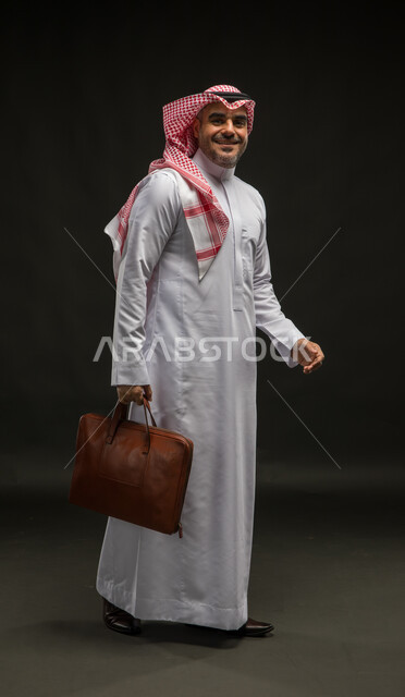 Feeling happy to achieve the company’s goals, going to work actively, portrait of a smiling Saudi Arabian Gulf man wearing a shemagh and traditional dress looking at the camera holding a handbag, interest in elegance and external appearance, full body image, black background