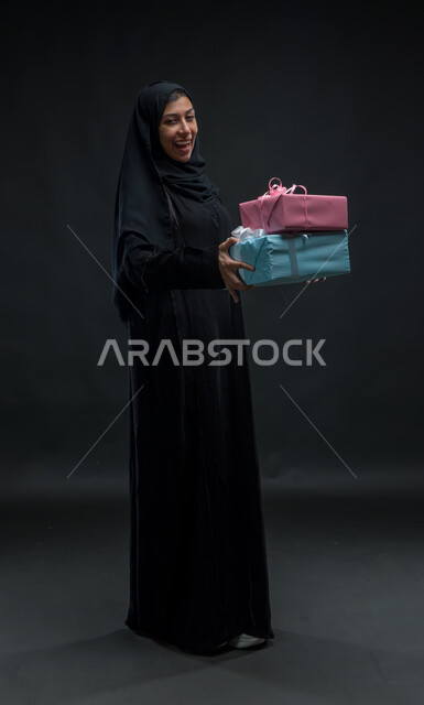  مناسبات و أعياد ومفاجآت سعيدة, إيماءات المفاجئة و السرور، فرحة الحصول على الهدية والتهنئة، النظر الى الكاميرا، بورتريه لامرأة عربية سعودية خليجية محجبة ترتدي عباءة سوداء تحمل بيديها علب مغلفة، صورة الجسم بالطول الكامل، خلفية سوداء