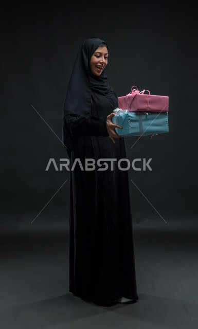 مفاجآت سعيدة و أعياد ومناسبات ,إيماءات الذهول و الاندهاش والفرح بالحصول على الهدية والتهنئة، بورتريه لامرأة عربية سعودية خليجية محجبة ترتدي عباءة سوداء تحمل بيديها علب هدايا مغلفة، صورة الجسم بالطول الكامل، خلفية سوداء