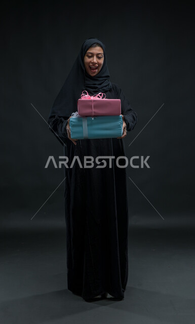 إيماءات المفاجئة و السرور، فرحة الحصول على الهدية والتهنئة، أعياد ومناسبات ومفاجآت سعيدة، بورتريه لامرأة عربية سعودية خليجية محجبة ترتدي عباءة سوداء تحمل بيديها علب مغلفة، صورة الجسم بالطول الكامل، خلفية سوداء