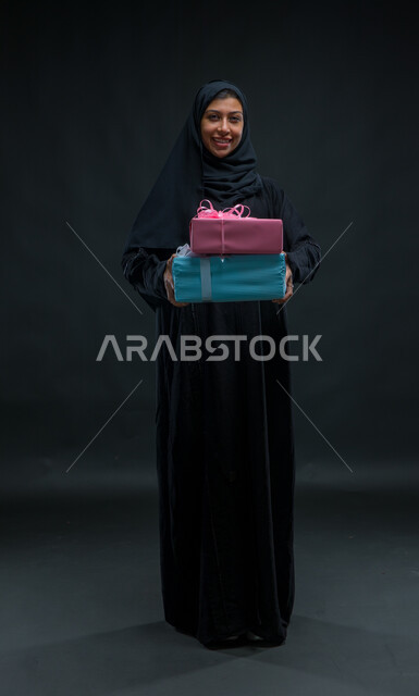 فرحة الحصول على الهدية والتهنئة، أعياد ومناسبات ومفاجآت سعيدة, النظر الى الكاميرا بإيماءات السرور، بورتريه لامرأة عربية سعودية خليجية محجبة ترتدي عباءة سوداء تحمل بيديها علب مغلفة، صورة الجسم بالطول الكامل، خلفية سوداء