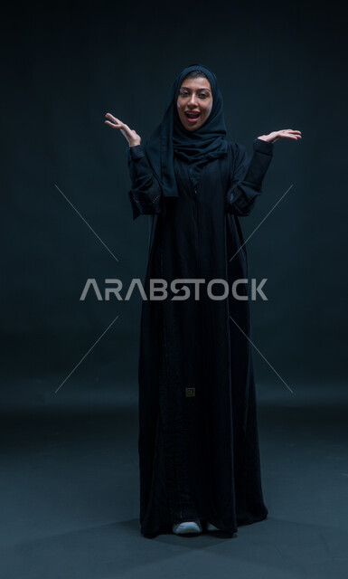 النظر إلى الكاميرا، قضاء اوقات ممتعه بالتسلية والترفيه ,ايماءات الدهشة والمفاجئة عند رؤية شيء ما، بورتريه لامرأة عربيه خليجية سعوديه ترفع يديها للاعلى بإشارات تدل على المفاجأة و اللعب ,ممارسة الالعاب الحركية ، خلفية سوداء