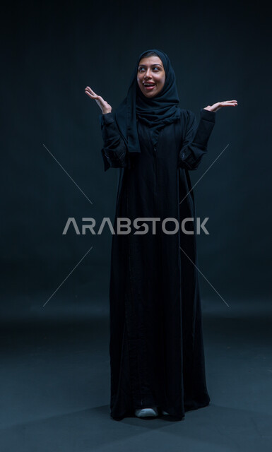 Surprising and surprising gestures when seeing something, spending fun times with entertainment and entertainment, portrait for a Saudi Gulf Arab woman who raises her hands to the top with signals indicating surprise and play, playing kinetic games, black background