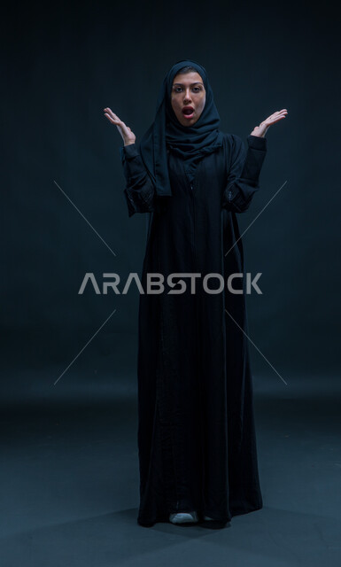 النظر إلى الكاميرا بإيماءات الذعر والخوف ,الشعور بالصدمة و الدهشة والتعجب والذهول , بورتريه لشابة عربية خليجية سعودية ترتدي العباءة و ترفع يديها للأعلى بتعابير التفاجؤ عند رؤية شيء ما, صورة الجسم بالطول الكامل, خلفية سوداء