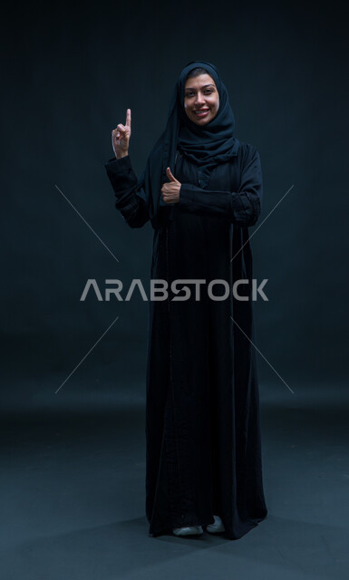 تعابير علامة الجودة، الاشارة إلى شيء ما، بورتريه لامرأة عربيه خليجيه سعودية ترتدي العباءه السوداء تقف باستقامة وتنظر إلى الكاميرا بإيماءات السرور، رفع الاصابع إلى الأعلى لتحديد مكان معين، صورة الجسم بالطول الكامل ، خلفية سوداء