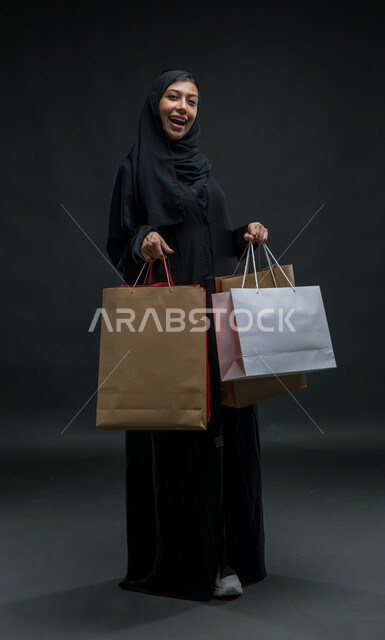  النظر إلى الكاميرا بتعابير الحماس للتسوق واقتناء المستلزمات والهدايا، ماركات عالمية فاخرة، عروض وتخفيضات موسمية في المولات، بورتريه لامرأة عربية خليجية سعودية محجبة ترتدي عباءة سوداء تحمل بيدها أكياس ورقية ملونة ، صورة للجسم بالكامل، خلفية سوداء