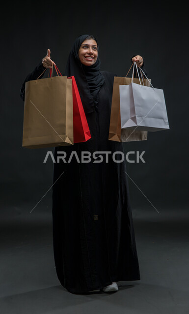 النظر للأعلى بتعابير الحماس للتسوق واقتناء المستلزمات والهدايا، إيماءات السرور والسعاده ،الاستفادة من عروض وتخفيضات الجمعة البيضاء، بورتريه لامرأة عربية خليجية سعودية محجبة ترتدي عباءة سوداء تحمل بيدها أكياس ورقية ملونة، صورة للجسم بالكامل، خلفية سوداء