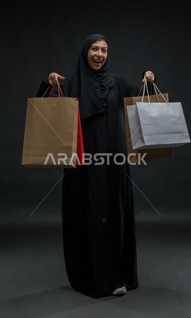 الاستفادة من عروض وتخفيضات الجمعة البيضاء، النظر إلى الكاميرا بتعابير الحماس للتسوق واقتناء المستلزمات والهدايا، بورتريه لامرأة عربية خليجية سعودية محجبة ترتدي عباءة سوداء تحمل بيدها أكياس ورقية ملونة بإيماءات السرور والسعادة، صورة للجسم بالكامل، خلفية سوداء