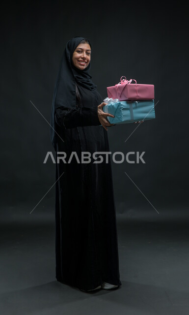 النظر الى الكاميرا بتعابير الفرح، مناسبات و أعياد ومفاجآت سعيدة, إيماءات المفاجئة و السرور، فرحة الحصول على الهدية والتهنئة، بورتريه لامرأة عربية سعودية خليجية محجبة ترتدي عباءة سوداء تحمل بيديها علب مغلفة، صورة الجسم بالطول الكامل، خلفية سوداء