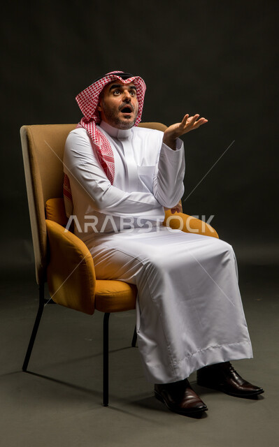 الاستفسار والاستفهام عن شيء ما، الاهتمام بالأناقة والمظهر الخارجي، بورتريه لرجل عربي خليجي سعودي مبتسم يرتدي الشماغ والثوب التقليدي يجلس على كرسي مريح ينظر إلى الأعلى بإيماءات الاندهاش، السؤال عن أمر معين بتعابير الاستغراب، خلفية سوداء.