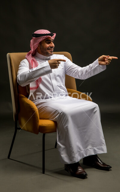 الإشارة إلى شيء ما، الاهتمام بالأناقة والمظهر الخارجي، بورتريه لرجل عربي خليجي سعودي مبتسم يرتدي الشماغ والثوب التقليدي يجلس على كرسي مريح ينظر إلى الكاميرا بإيماءات الفرح، مفهوم الرجولة و الطموح والثقة بالنفس، خلفية سوداء.