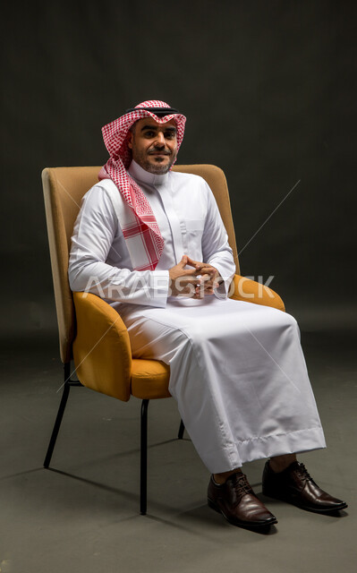 The concept of masculinity, ambition and self-confidence, interest in elegance and external appearance, a portrait of a smiling Saudi Gulf Arab man wearing a shemagh and traditional thobe, sitting on a comfortable chair looking at the camera with happy gestures, black background