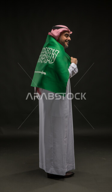 إيماءات الفرح في يوم العلم  11 مارس، الشعور بالحماس في اليوم الوطني السعودي 23 سبتمبر، بورتريه من الخلف لرجل عربي خليجي سعودي يرتدي الشماغ والثوب التقليدي يقف باستقامة يضع راية المملكة على اكتافه، صورة الجسم بالطول الكامل, خلفية سوداء