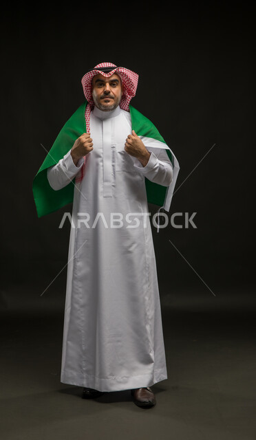 الشعور بالحماس في اليوم الوطني السعودي 23 سبتمبر، الوقوف باستقامة والنظر إلى الكاميرا بإيماءات الفرح في يوم العلم  11 مارس، بورتريه لرجل عربي خليجي سعودي يرتدي الشماغ والثوب التقليدي يضع راية المملكة على اكتافه، صورة الجسم بالطول الكامل, خلفية سوداء