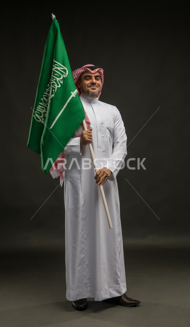 الوقوف باستقامة والنظر إلى الكاميرا بإيماءات الفرح في يوم العلم  11 مارس، مشاعر الحماس في الاحتفال باليوم الوطني السعودي 23 سبتمبر، بورتريه لرجل عربي خليجي سعودي يرتدي الشماغ والثوب التقليدي يحمل بيده راية المملكة، صورة الجسم بالطول الكامل, خلفية سوداء