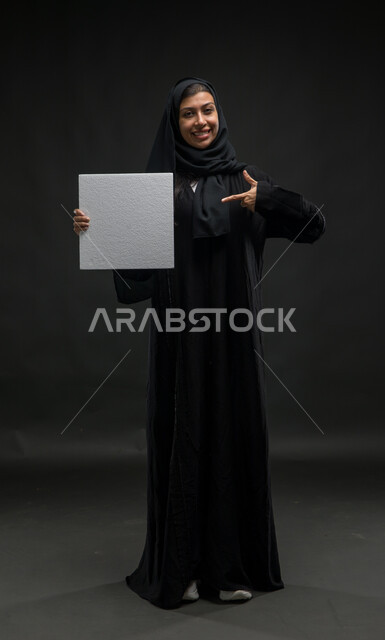 استخدام لوحات تعريفية توضيحيه، الوقوف باستقامة والاشارة الى لوحة بيضاء مربعة فارغة، إعلانات ودعايات وعروض تسويقية، بورتريه لإمرأة عربية خليجية سعودية مبتسمة محجبه ترتدي عباءة سوداء تحمل بيدها موك اب مربع أبيض، صورة للجسم بالكامل، خلفية سوداء