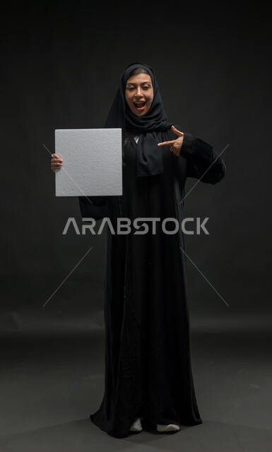 لوحة بيضاء مربعة فارغة، ايماءات الحماس لعرض إعلانات ودعايات وعروض تسويقية، بورتريه لإمرأة عربية خليجية سعودية محجبة مبتسمة ترتدي عباءة سوداء و تشير باصبعها الى موك اب مربع أبيض، استخدام لوحات توضيحية تعريفية، صورة الجسم بالطول الكامل، خلفية سوداء