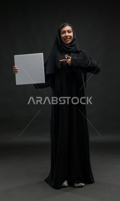 Standing with integrity and reference to an empty square white painting, advertisements, advertisements and marketing offers, portrait for a Saudi Gulf -Gulf woman smiling veiled wearing a black cloak carrying a white box makeup, using introductory illustrations, a picture of the entire body, black background