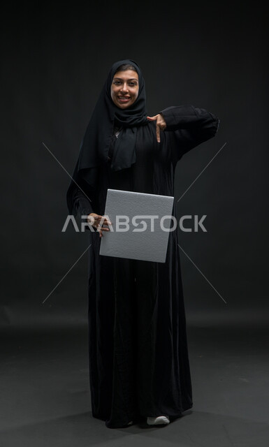 Using introductory illustrations, standing up and reference to an empty square white painting, advertisements, advertisements and marketing offers, portrait for a Saudi -Gulf Arab woman smiling with veiled wearing a black cloak carrying a white box of a white box, a picture of the entire body, black background
