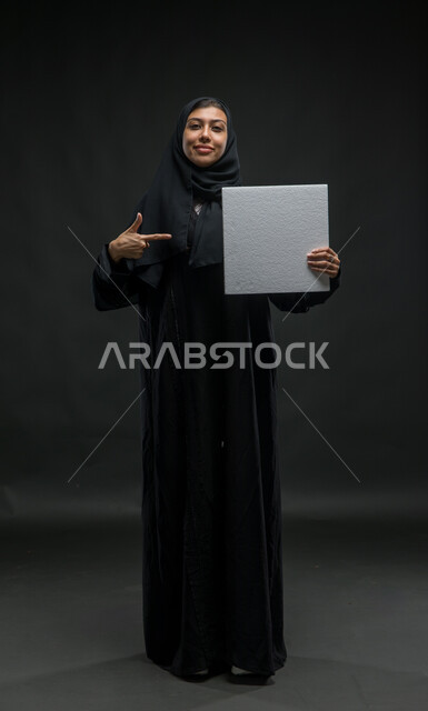 الوقوف باستقامة والاشارة الى  لوحة بيضاء مربعة فارغة، إعلانات ودعايات وعروض تسويقية، بورتريه لإمرأة عربية خليجية سعودية مبتسمة محجبه ترتدي عباءة سوداء تحمل بيدها موك اب مربع أبيض، استخدام لوحات توضيحية تعريفية، صورة للجسم بالكامل، خلفية سوداء