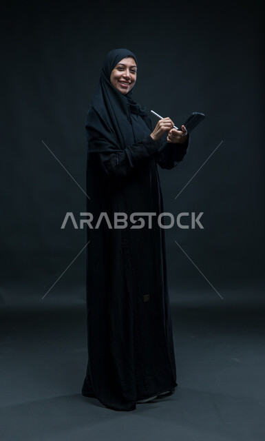 دمج التقنيات الحديثة والتكنولوجيا في الحياة العملية، النظر إلى الكاميرا بتعابير السرور, بورتريه لإمرأة عربية خليجية سعودية محجبة ترتدي العباءة السوداء تقوم باستخدام قلم الجهاز اللوحي لتسجيل وتدوين البيانات والملاحظات، صورة للجسم بالكامل، خلفية سوداء