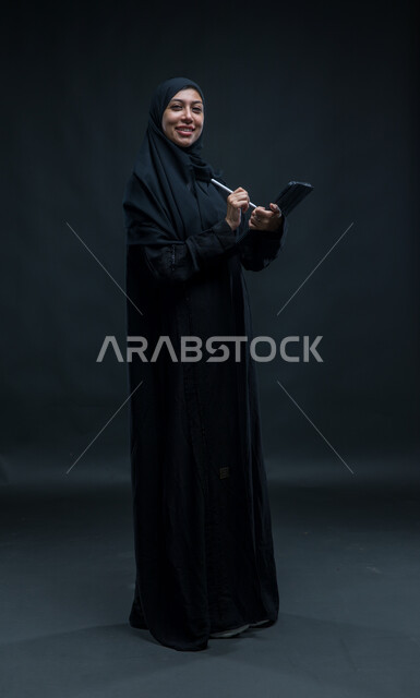 Completion of work tasks through the tablet, looking at the camera with pleasure expressions, modern technical devices,   Portrait for a veiled Arab Gulf Arab woman wearing a black cloak uses the tablet pen to record and record data and notes, a picture of the entire body, black background