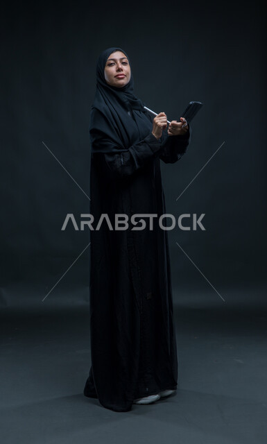 Looking at the camera with self -confidence expressions, using the tablet pen to record and record data and notes, modern technical devices, portrait for a veiled Arab Gulf Arab woman who wears the black cloak that accomplishes work tasks through the tablet, the entire body image, black background