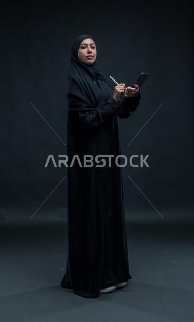Use the tablet pen to record and record data and notes, modern technical devices, portrait for a veiled Arab Gulf woman who wears the black cloak that accomplishes the work tasks through the tablet, looking at the camera with self -confidence expressions, the entire body image, black background
