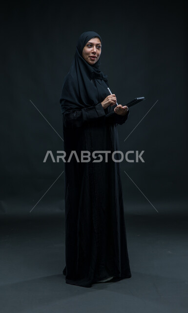 تعابير الاستفسار عن معلومات, استخدام قلم الجهاز اللوحي لتسجيل وتدوين البيانات والملاحظات، اجهزة تقنية حديثة، بورتريه لإمرأة عربية خليجية سعودية محجبة ترتدي العباءة السوداء تقوم بانجاز مهام العمل عبر جهاز التابلت, صورة الجسم بالكامل، خلفية سوداء