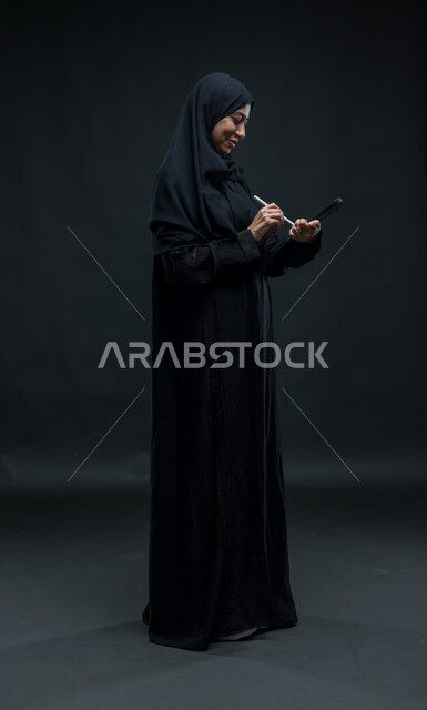 Expressions of pleasure and communication with one of the friends, the use of the tablet pen of the shield through the social media programs, modern technical devices, portrait for a veiled Arab Gulf Arab woman who wears the black cloak that accomplishes work tasks through the tablet, the entire body image, black background