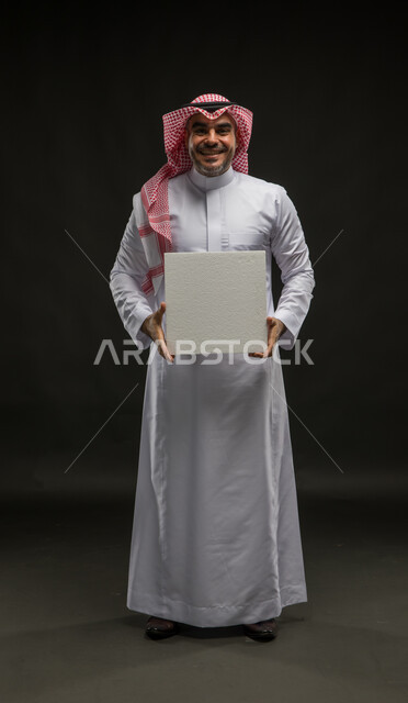 استخدام لوحات توضيحية ووسائل تعليمية، إعلانات وعروض تسويقية، موك اب مكعب أبيض، بورتريه لشاب عربي خليجي سعودي مبتسم يرتدي الشماغ والثوب التقليدي ينظر إلى الكاميرا يمسك بيده لوحه بيضاء مكعبه فارغة، إيماءات الحماس والفرح، صورة الجسم بالطول الكامل، خلفية سوداء