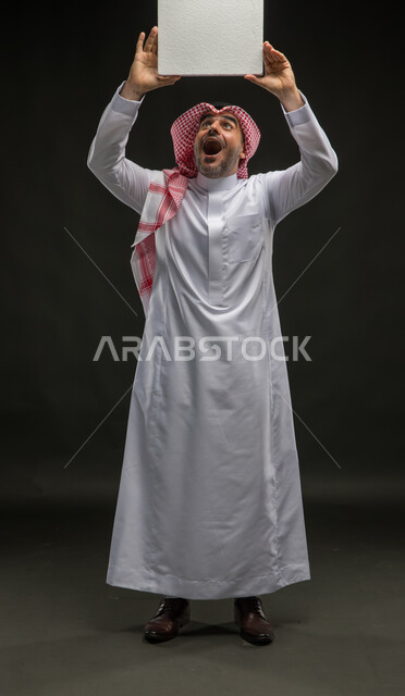 موك اب مكعب أبيض، إعلانات وعروض تسويقية، استخدام لوحات توضيحية ووسائل تعليمية، بورتريه لشاب عربي خليجي سعودي مبتسم يرتدي الشماغ والثوب التقليدي يرفع يديه إلى الأعلى يمسك لوحه بيضاء مكعبه فارغة ينظر إليها، إيماءات الذهول والاندهاش، صورة الجسم بالطول الكامل، خلفية سوداء