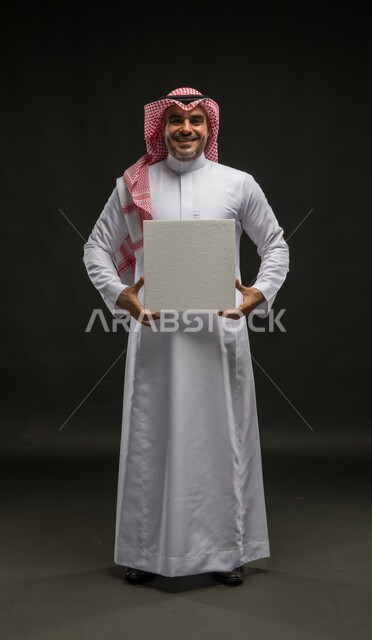 Advertisements and marketing presentations, a white cubic mockup, use of explanatory panels and teaching aids, a portrait of a smiling Saudi Gulf Arab young man wearing a shemagh and traditional dress, looking at the camera, holding a blank white cubic canvas in his hand, full-length body portrait, black background