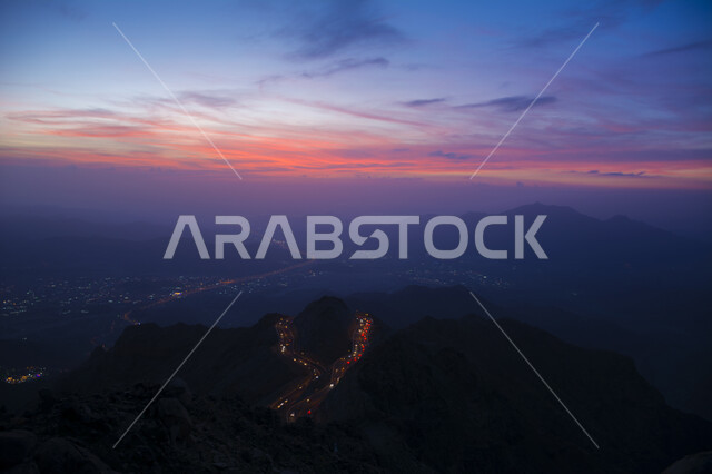 A sunset view over a bumpy, winding asphalt road in the middle of the rocky mountains in the Aqabat Al-Hada area, the mountainous environment, peaks and heights in the Kingdom of Saudi Arabia, an aerial photo from above of traffic and movement of cars on the Al-Hada Mountains in the city of Taif