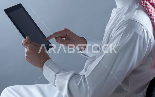 مفهوم التسوق والشراء اونلاين، إيماءات النقر والضغط على الجهاز اللوحي, بورتريه مقرب لرجل عربي خليجي سعودي يرتدي الشماغ والثوب التقليدي يجلس على الكرسي يحمل بيده التابليت، انجاز الاعمال والمهام عن بعد، خلفية بيضاء