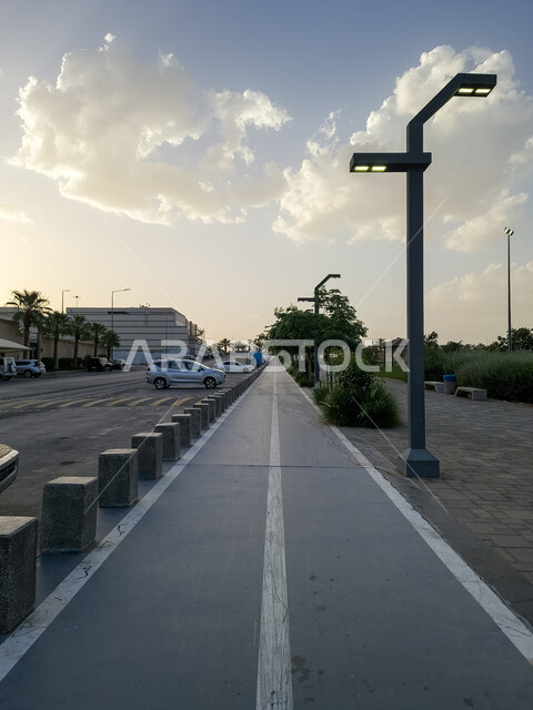 ممر اسفلتي في إحدى حرائق مدينة الرياض، التنزه في المنتزهات العامة في مدينة الرياض، منظر السماء المليئة بالغيوم، أعمدة إنارة الطرق والشوارع، زراعة الأشجار والنباتات الخضراء في المدن