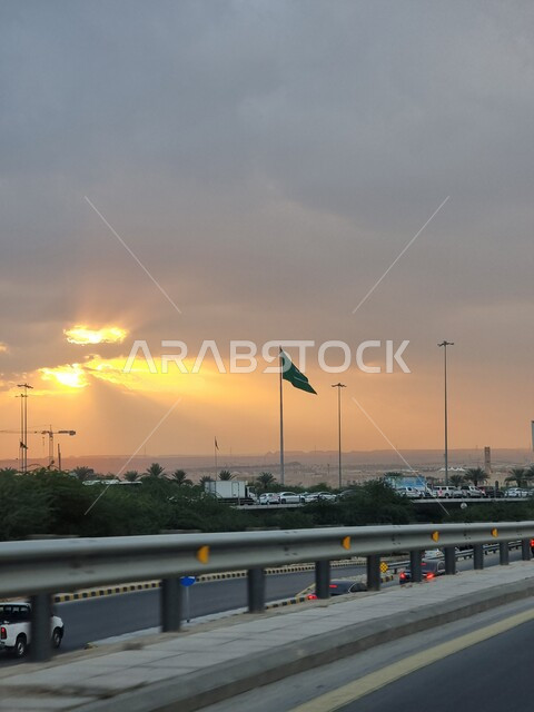 العلم الوطني السعودي يرفرف في سماء المملكة العربية السعودية في وقت غروب الشمس، راية التوحيد والإسلام، الاهتمام بزراعة الأشجار والنباتات الخضراء في المدن، حركة مرور السيارات في الشوارع والطرق الاسفلتية