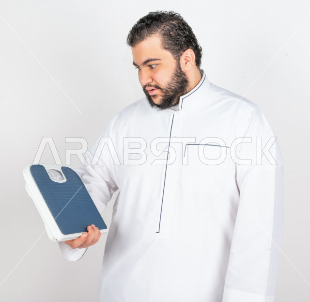 بورتريه شاب سعودي خليجي ممتلئ الجسم ، يعاني من السمنة  ،   مع ميزان لقياس الوزن ،مشاكل صحية ،الوزن الزائد والسمنة ، تراكم السعرات الحرارية 