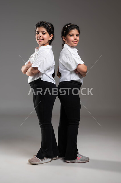 المساندة والدعم الاخوي، الاناقة والاهتمام بالمظهر الخارجي، مفهوم التوأم الحقيقي المتطابق، بورتريه لتوأم بنات عربيات خليجيات سعوديات مبتسمات، الوقوف باستقامة وأيدي متقاطعة والنظر الى الكاميرا بايماءات وتعابير الثقة بالنفس، صورة الجسم بالكامل، خلفية رمادية