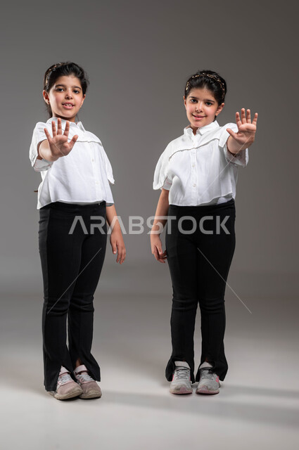 التعبير عن الرفض والإمتناع، إيماءات يدين تدل على الأمر بالتوقف عن فعل شيء ما, طلب عدم تخطي الحدود، بورتريه لتوأم بنات عربيات خليجيات سعوديات يوقومون بوضع كفيهما للأمام، صورة الجسم بالطول الكامل، خلفية رمادية
