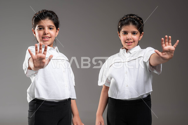 إيماءات يدين تدل على الأمر بالتوقف عن فعل شيء ما, التعبير عن الرفض والإمتناع، طلب عدم تخطي الحدود، بورتريه مقرب لتوأم بنات عربيات خليجيات سعوديات يوقومون بوضع كفيهما للأمام، خلفية رمادية