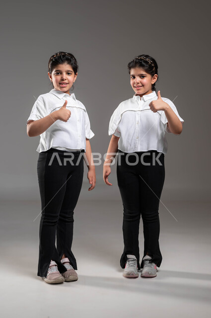 القبول والرضا بالنتيجة، ايماءات الاعجاب والموافقة، بورتريه لأختين توأم عربيتين خليجيتين سعوديتين ترتديان الملابس الكاجوال وترفعان اصبع الابهام للأعلى، الاتفاق على أمر معين، صورة الجسم بالطول الكامل، خلفية رمادية