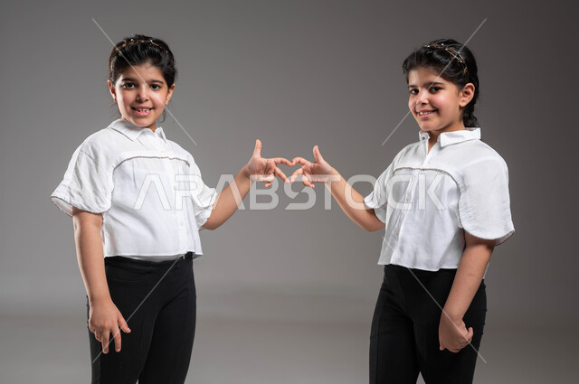 المحبة والمودة بين الشقيقات، بورتريه مقرب لأختين توأم عربيتين خليجيتين سعوديتين مبتسمتين ترتديان الملابس الكاجوال وتشكلن قلب حب بيديهما تنظران الى الكاميرا بتعابير السرور، تقوية وتعزيز الروابط العائلية، معنى الصداقة والاخوة، خلفية رمادية