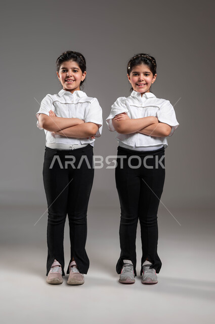 The concept of real identical twins, wearing casual clothes and paying attention to the external appearance, a portrait of twin smiling Saudi Gulf Arab girls, standing straight with crossed hands and looking at the camera with gestures and expressions of self-confidence, full body portrait, gray background